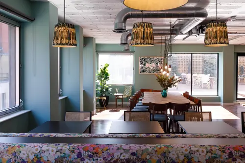 Breakout space at Creo London Chancery Lane with a long communal table, cane-backed chairs, patterned pendant shades, exposed ductwork and windows onto a terrace.