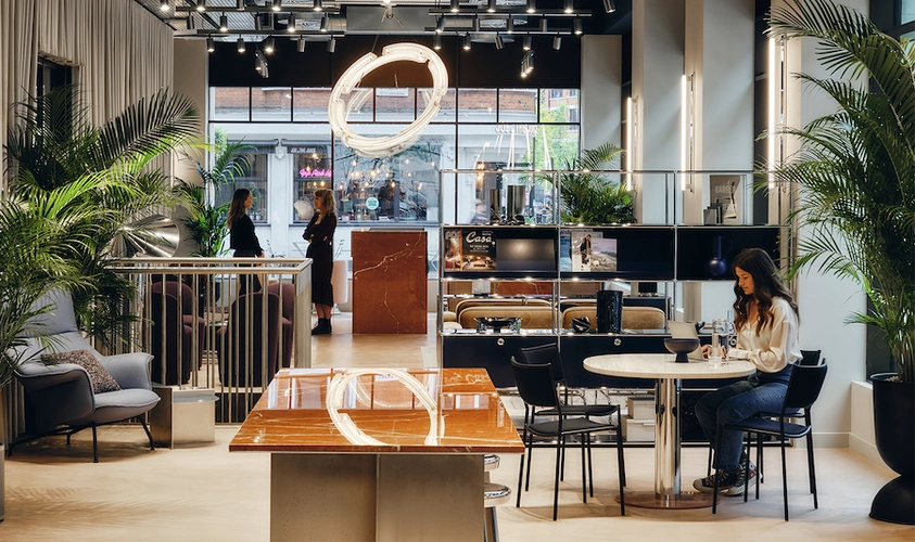 Gallery lounge at FORA Brock House filled with loads of lights and green plants with a woman coworking at the table and two women facing each other standing in the background