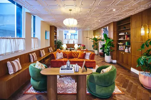 Lounge area at Creo Bank, 1 Royal Exchange Avenue with orange velvet sofas, green armchairs, a built-in wooden bench and shelving, herringbone wood floor and an ornate moulded ceiling with pendant lanterns.